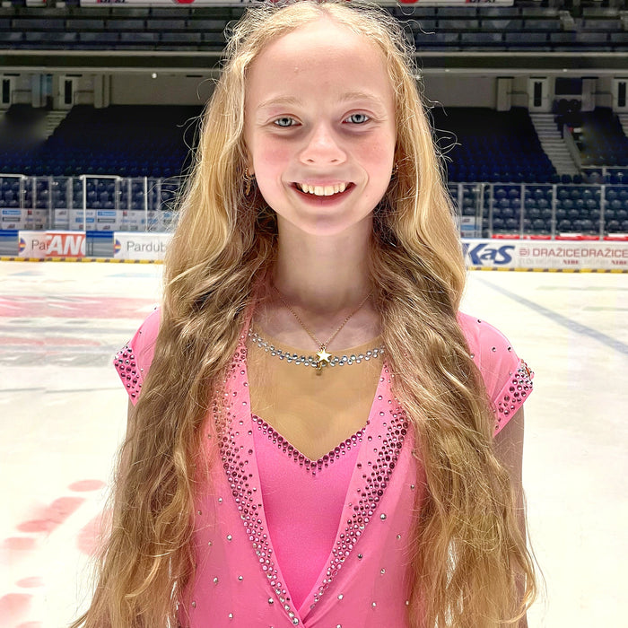 figure skater standing on ice in a pink sparkly dress wearing a gold necklace