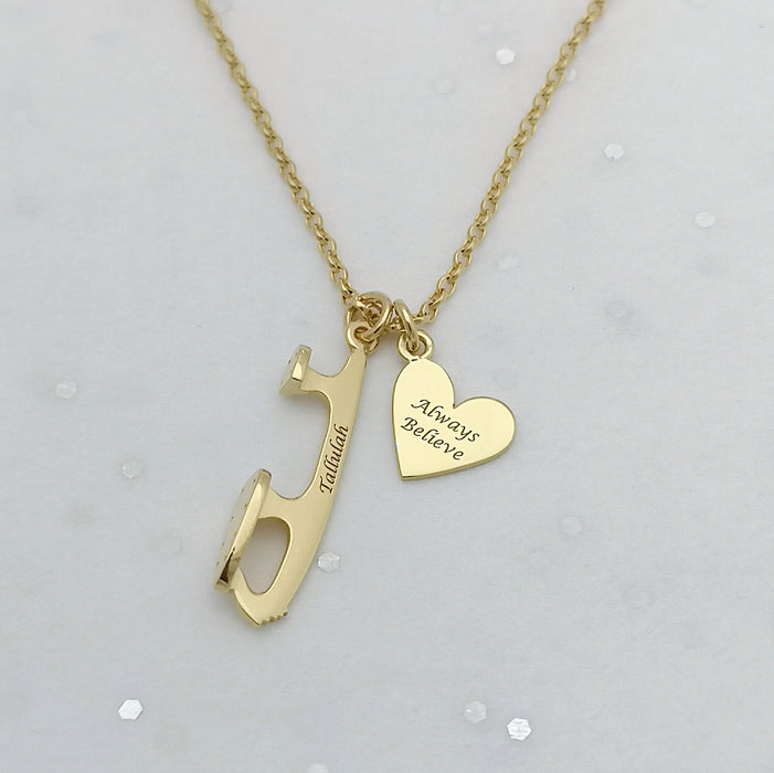 A gold-colored necklace featuring a custom ice skating blade pendant with the word 'BELIEVE' and a heart shape on a chain.