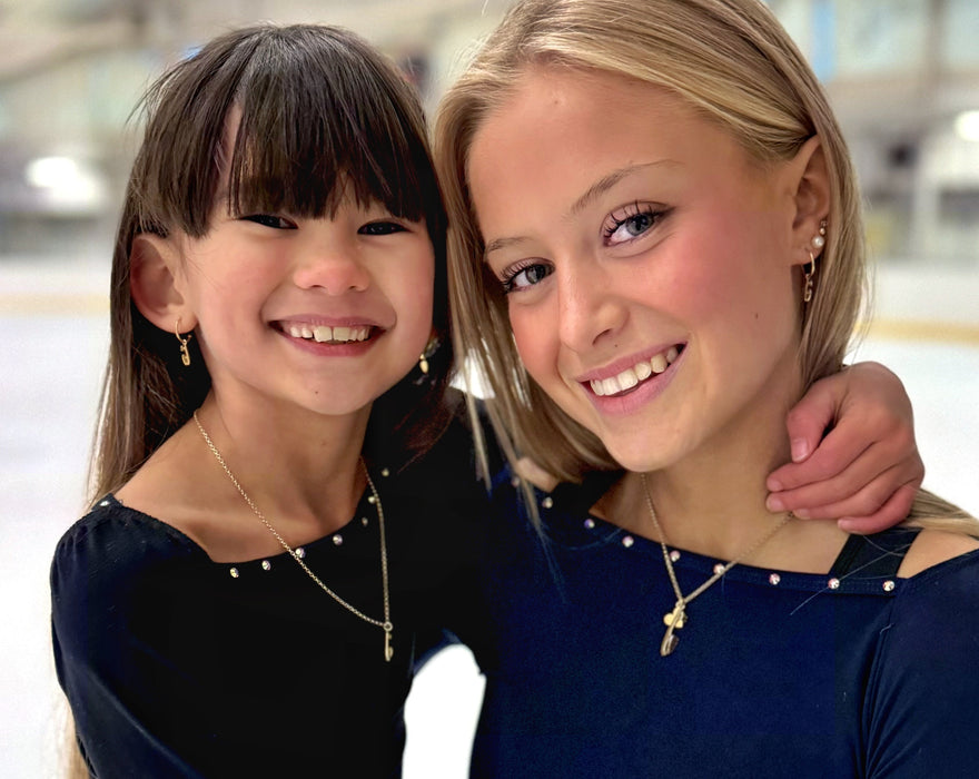 Close up of two figure skaters in dark blue and black tops on an ice rink wearing gold ice skate blade necklaces