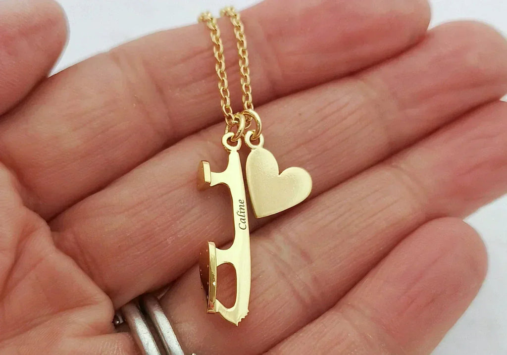 close up of a hand holding a gold plated necklace with an engraved ice skate blade charm and a heart charm