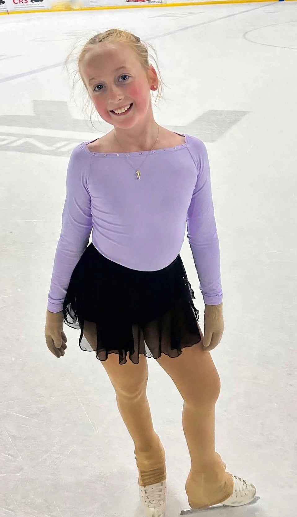 figure skater standing on an ice rink in a purple top and black skirt wearing an ice skate blade necklace