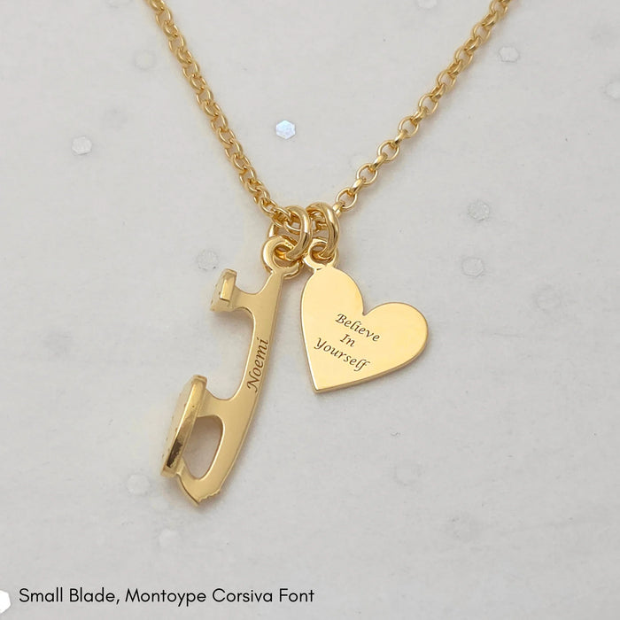 Gold necklace with a heart and an ice skate blade charm, both charms are engraved on a pale grey background