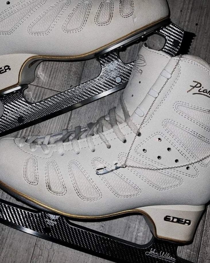 White figure skating boots with black blades on a wooden floor, draped on top of the boots is a silver ice skate blade necklace