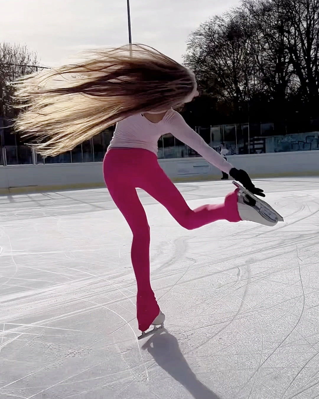 a figure skater on an outdoor ice rink wearing pink leggings, she is catching her foot to practice a beilmann spin
