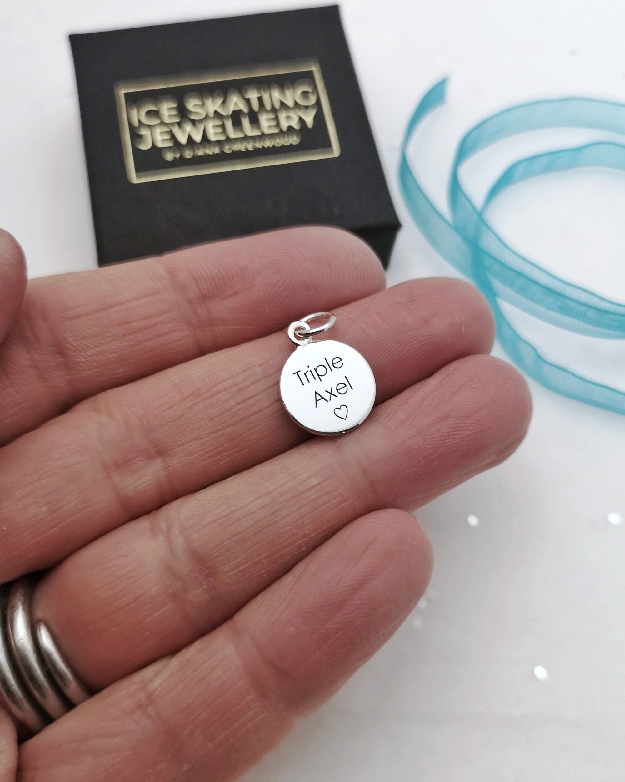 hand holding an engraved silver disc charm that reads Triple Axel, a jewellery box is visible in the background that reads Ice Skating Jewellery