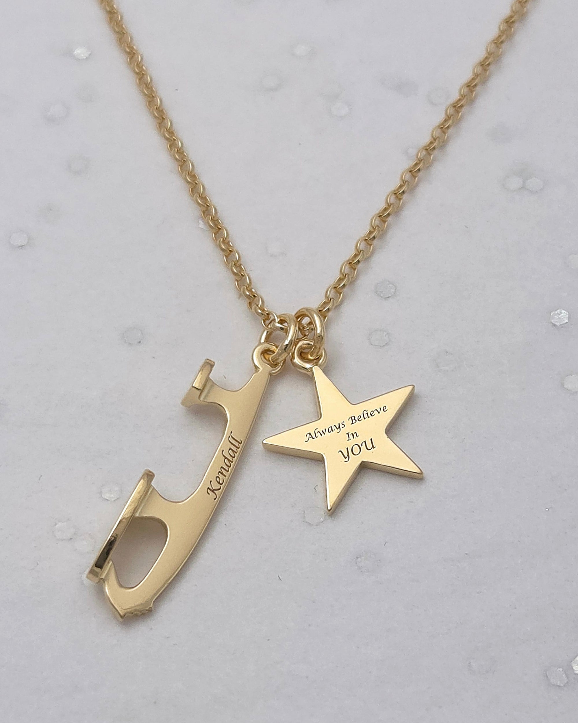 Gold skating necklace with two engraved charms — ice skate blade with ‘Kendall’ and star charm reading ‘Always Believe In You’ on grey background.