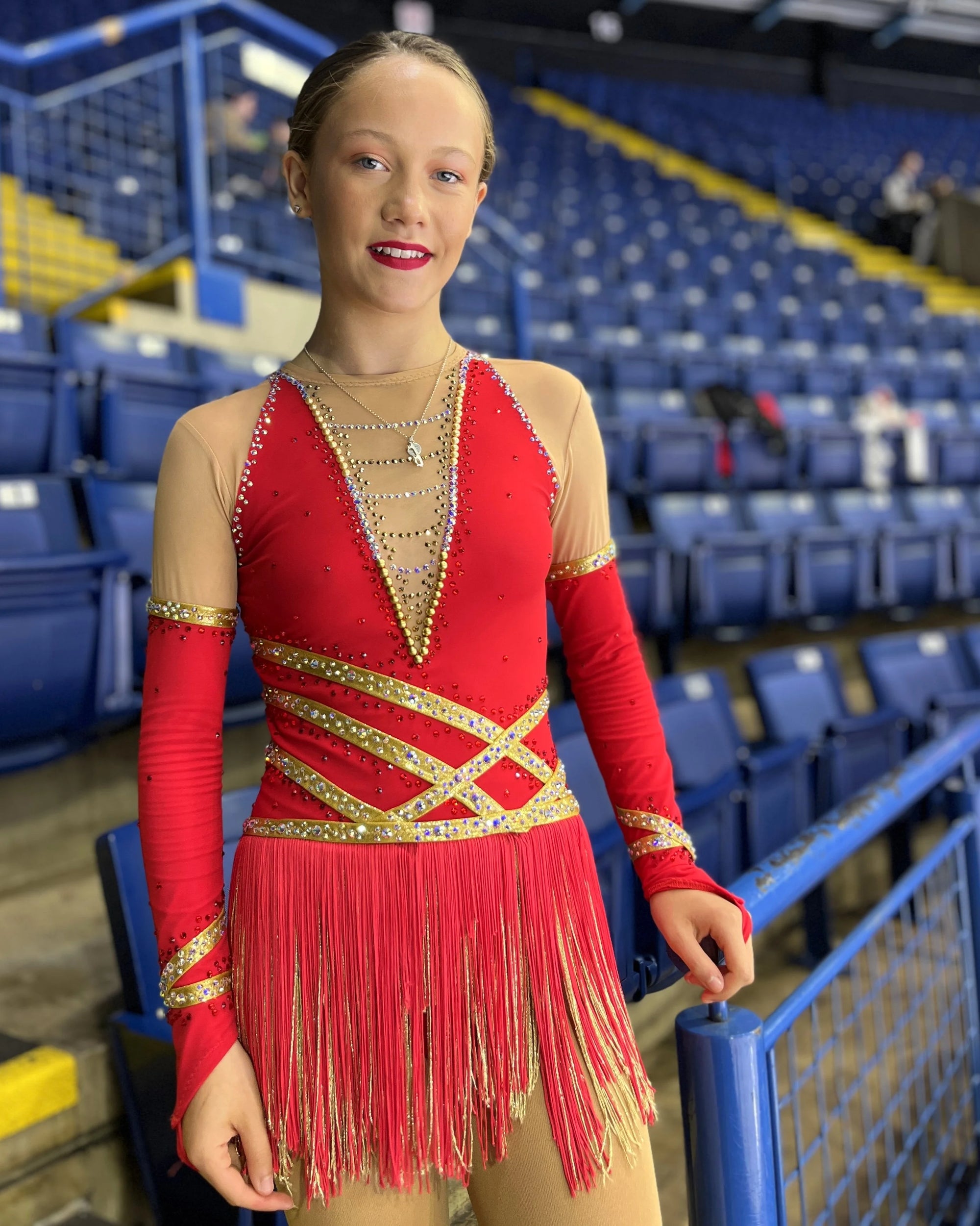a figure skaterstanding ready to perform in a red sparkly dress and red lipstick with the blue seats of the ice arena behind her