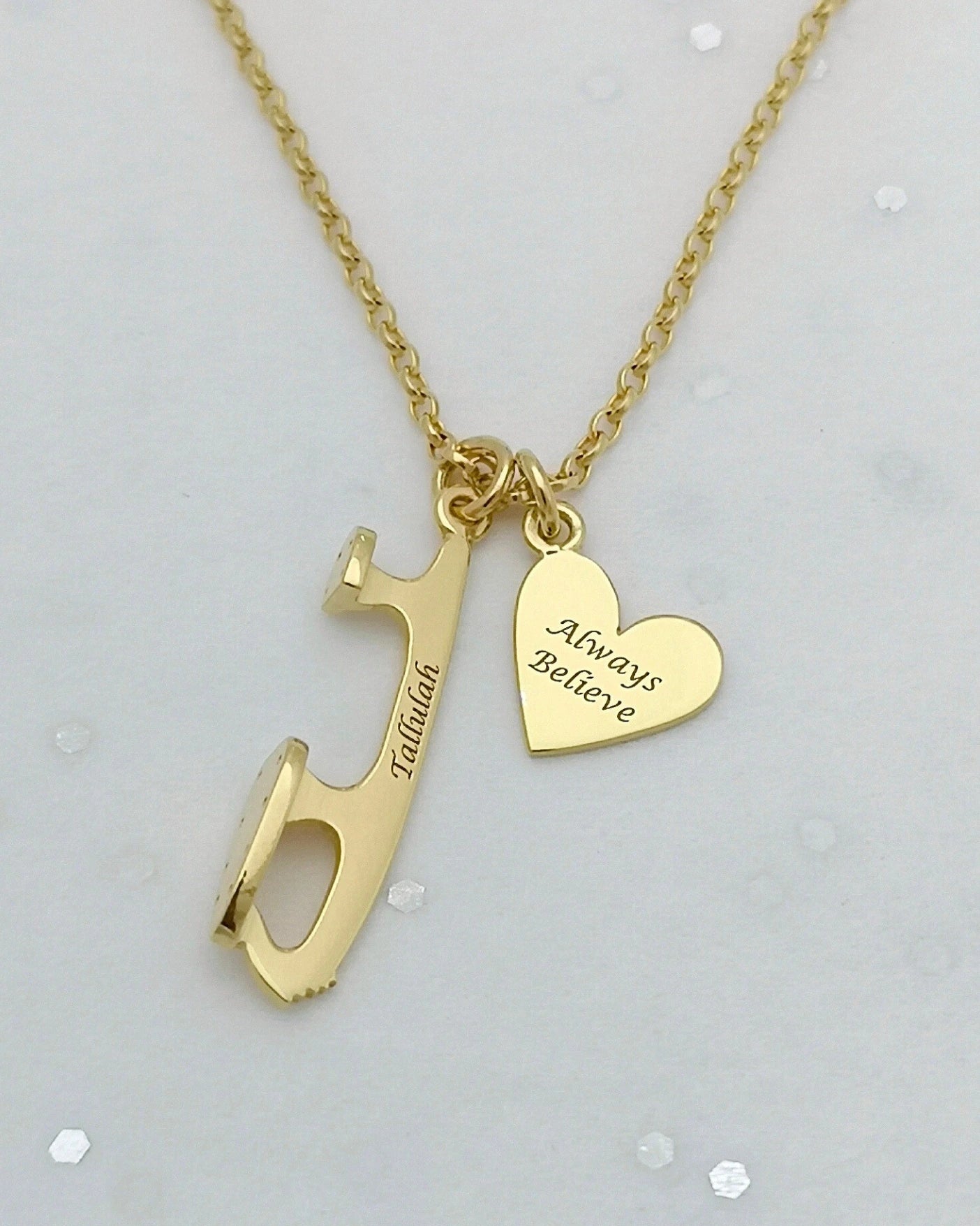 gold necklace with two engraved charms - an ice skate blade charm that reads Tallulah and a heart that says Always Believe