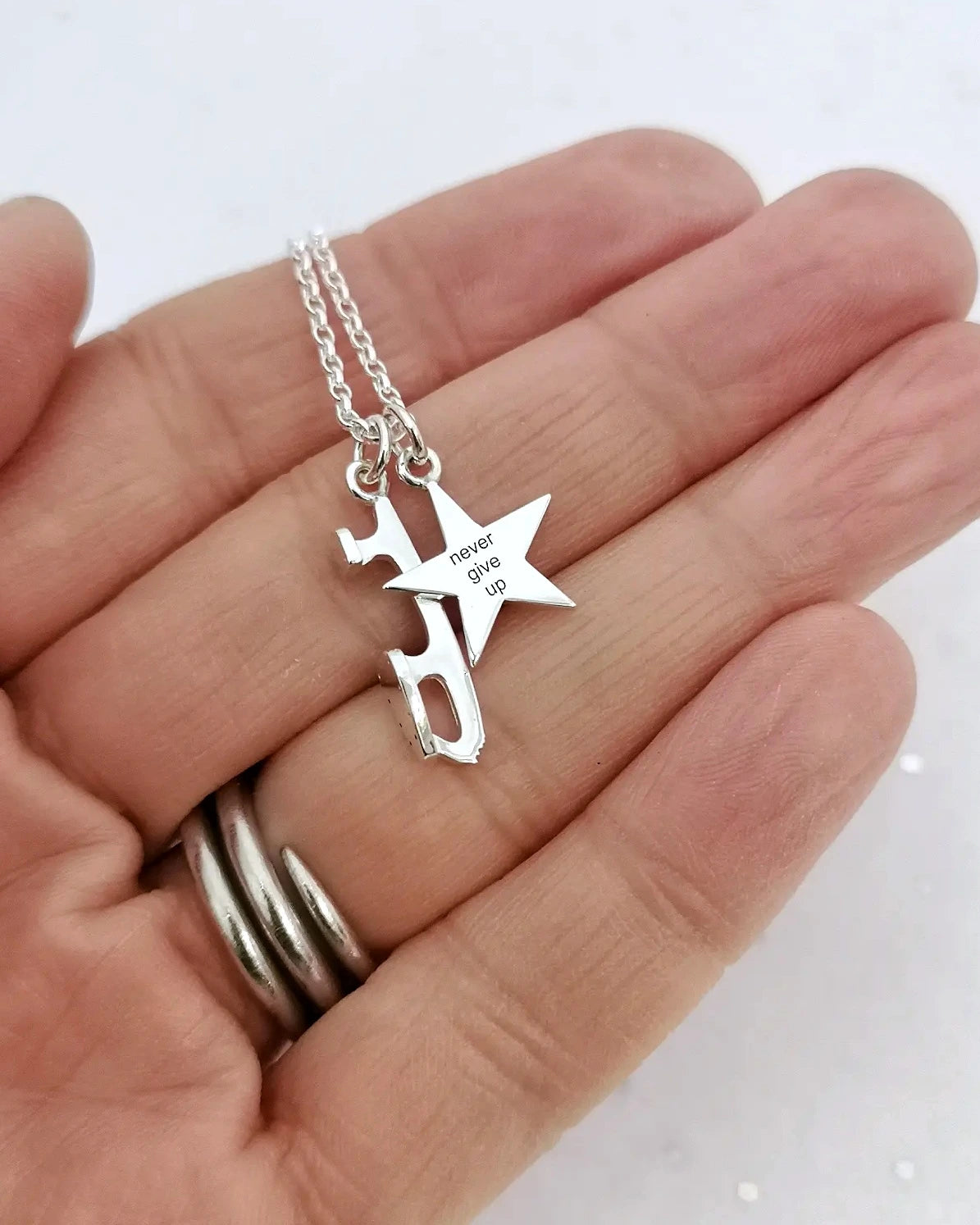 close-up of a hand holding a silver necklace with two charms - an ice skate blade and an engraved star which says 'never give up' 