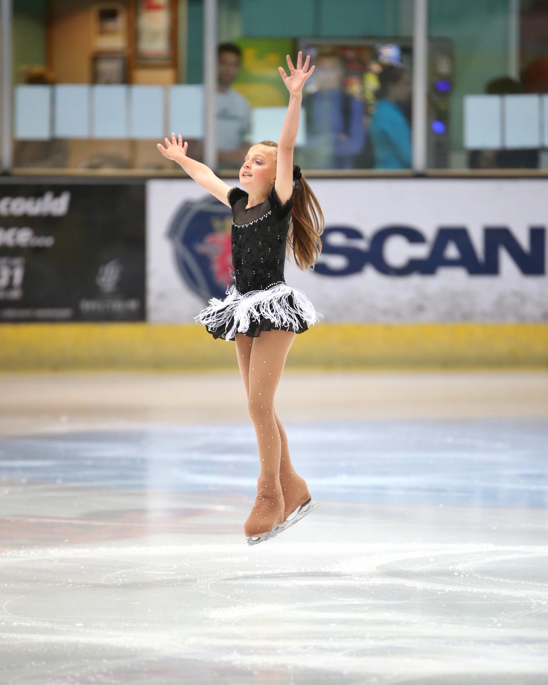 Young figure skater captured mid-air during routine with outstretched arms and tasselled skirt in motion.