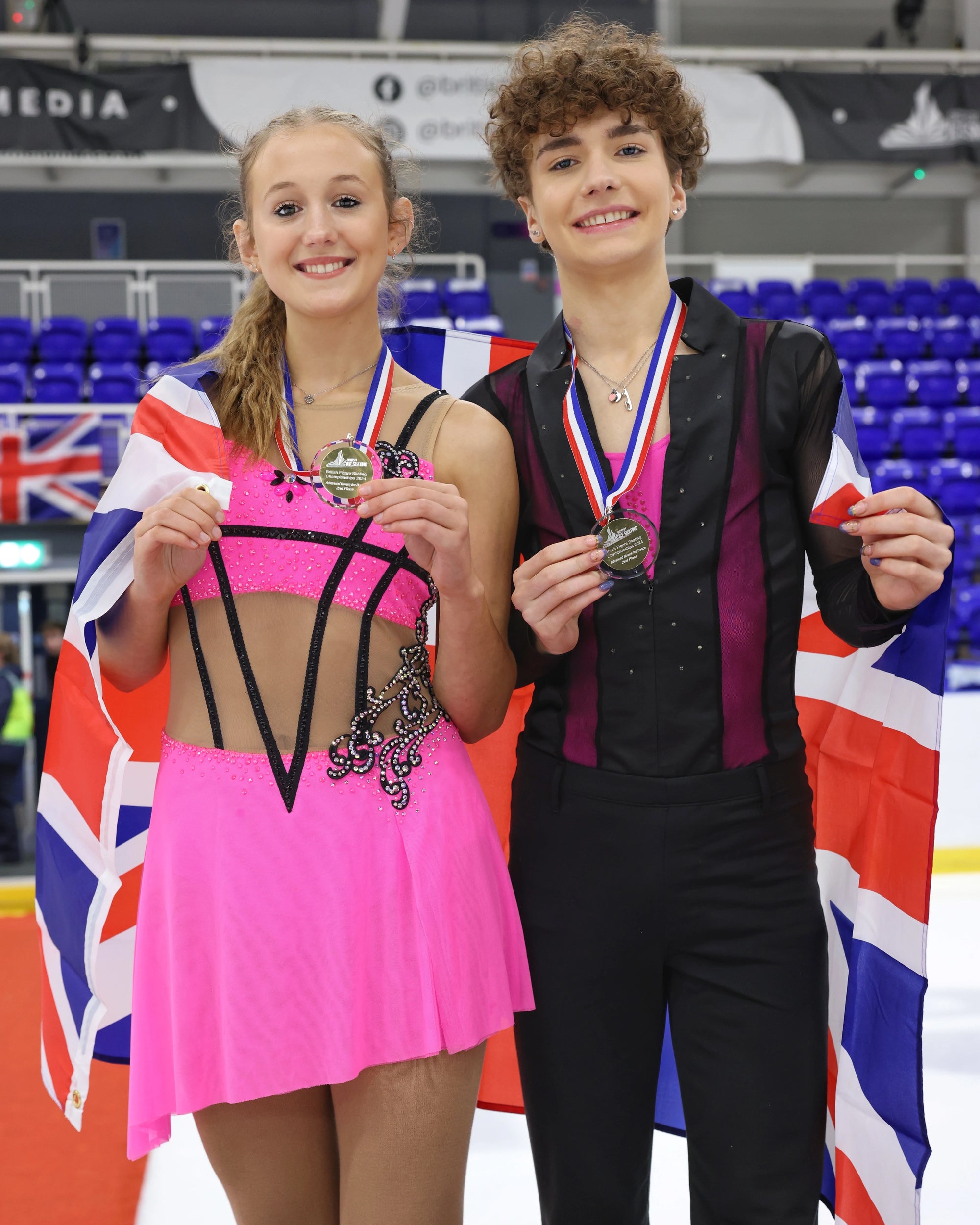 two figure skater standing on a podium wearing their sparkly costumes and holding their winners medals 