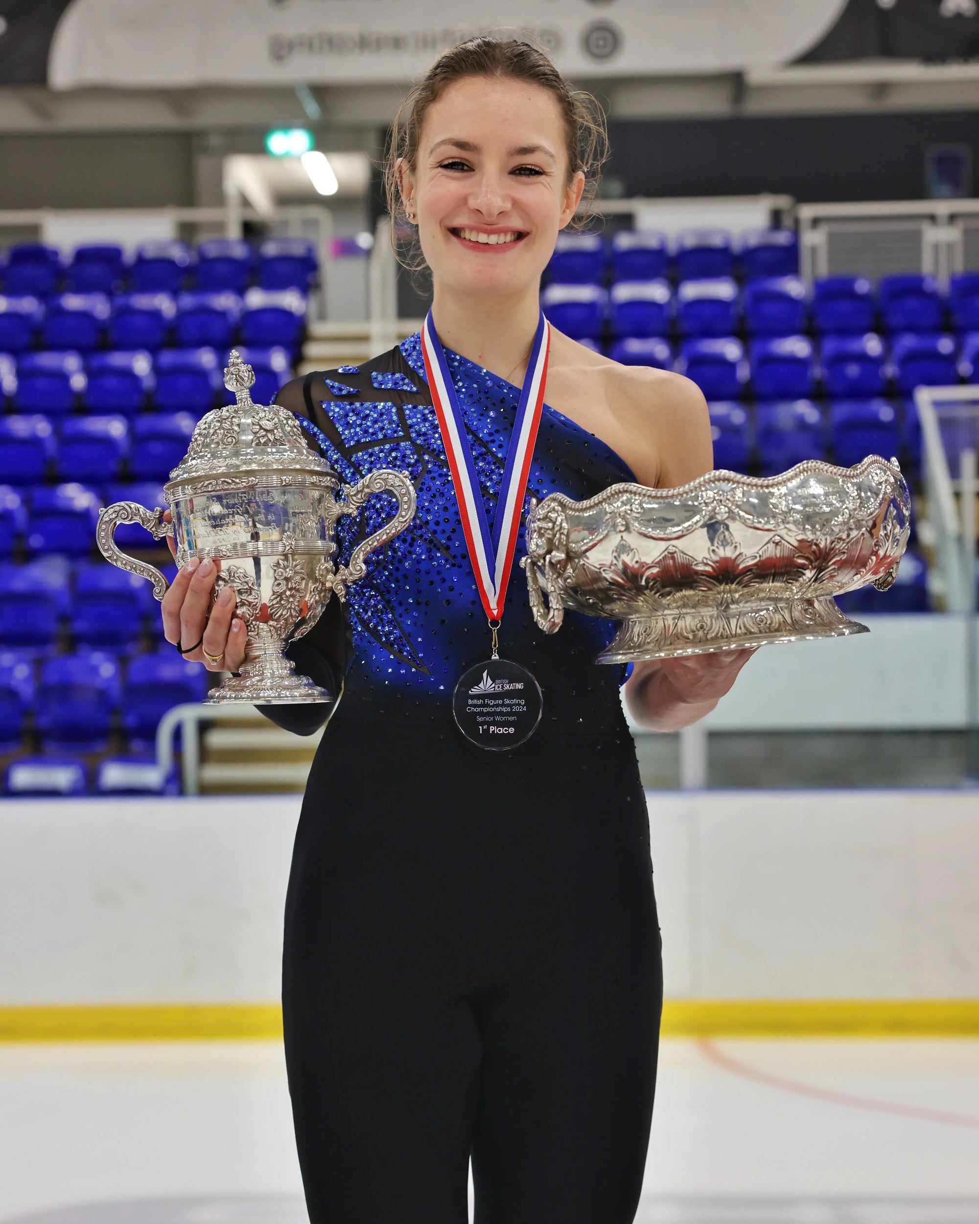 Kristen Spours standing on podium on ice wearing sparkly costume, holding two trophies after winning British Championship.