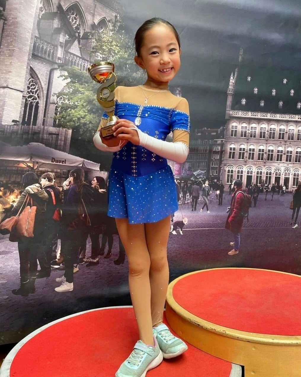 young skater in a blue sparkly costume on a podium holding her winning trophy and smiling