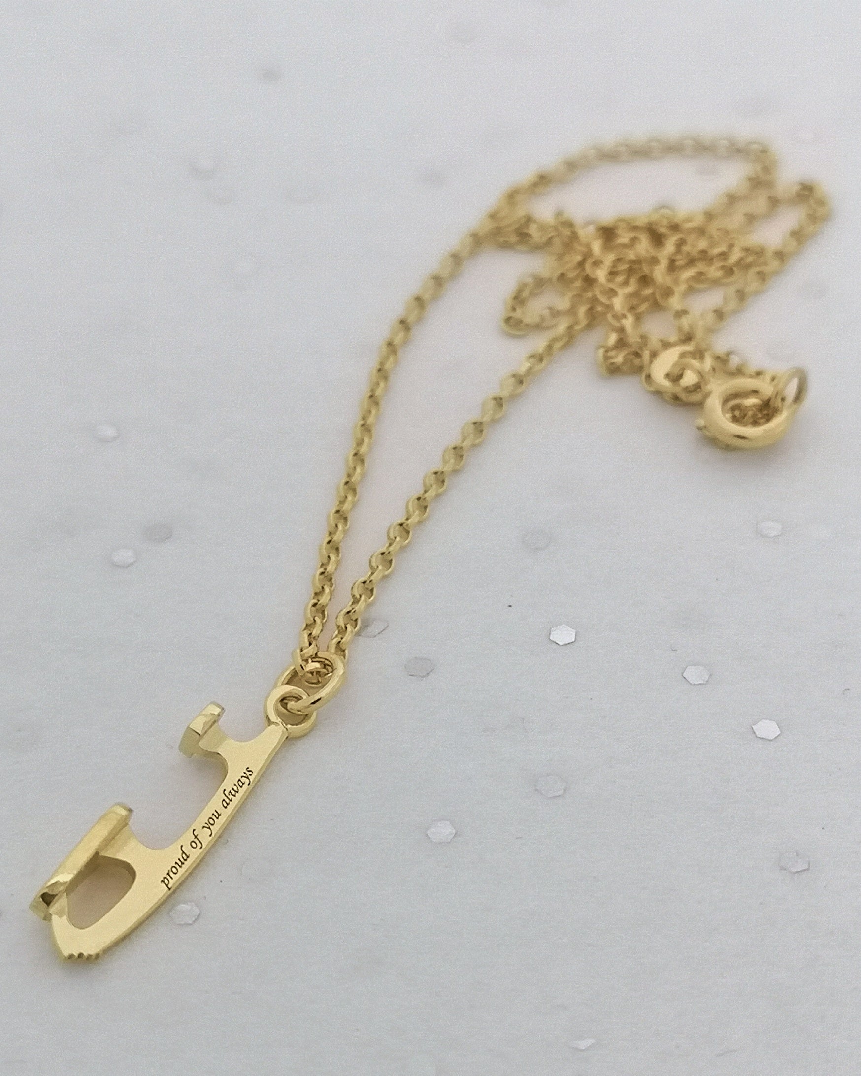 close-up of a gold ice skating necklace with the words 'proud of you always' engraved along on the tiny ice skating blade