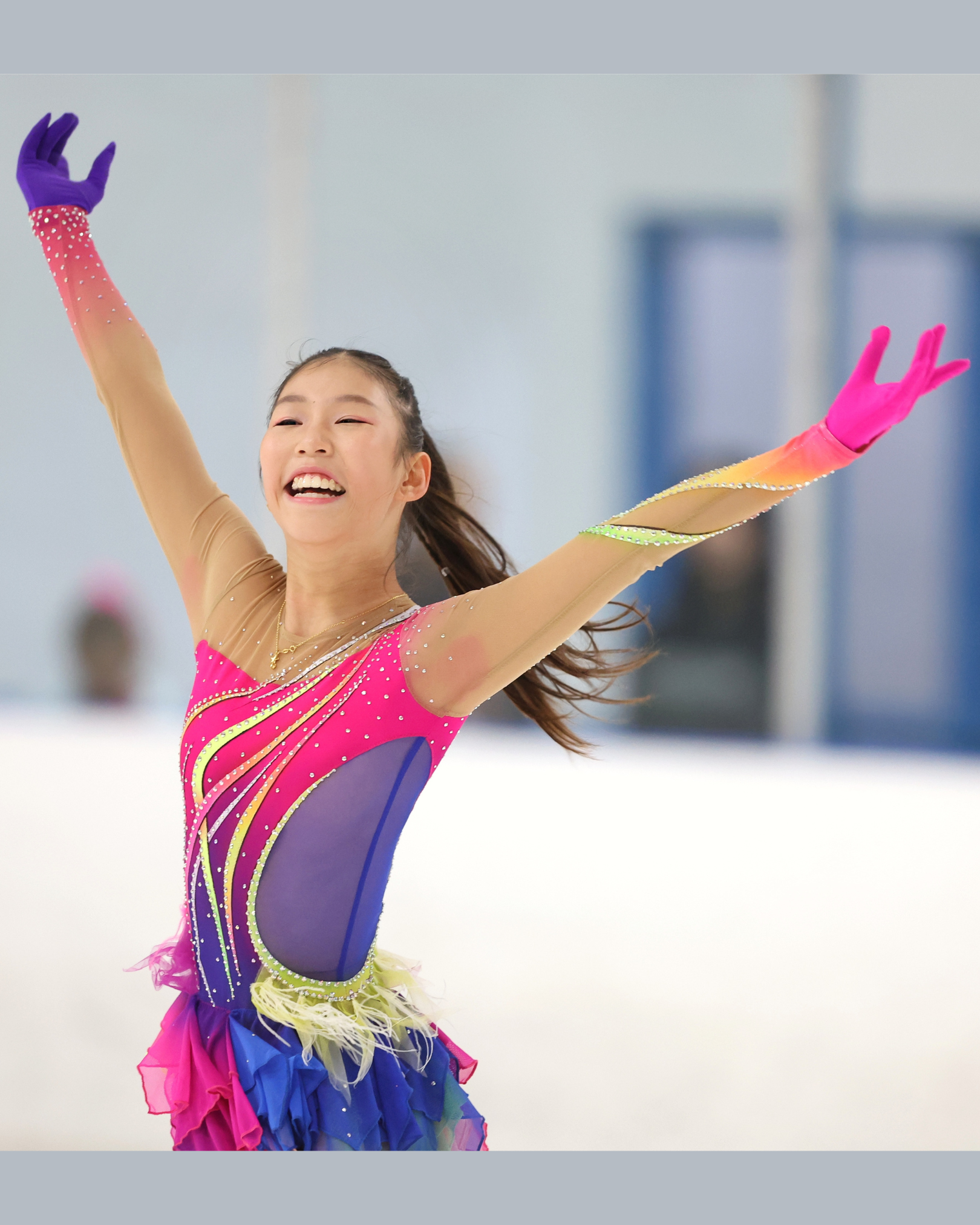 Figure skater in bright sparkly dress on ice with arms outstretched, smiling joyfully at the end of her programme.