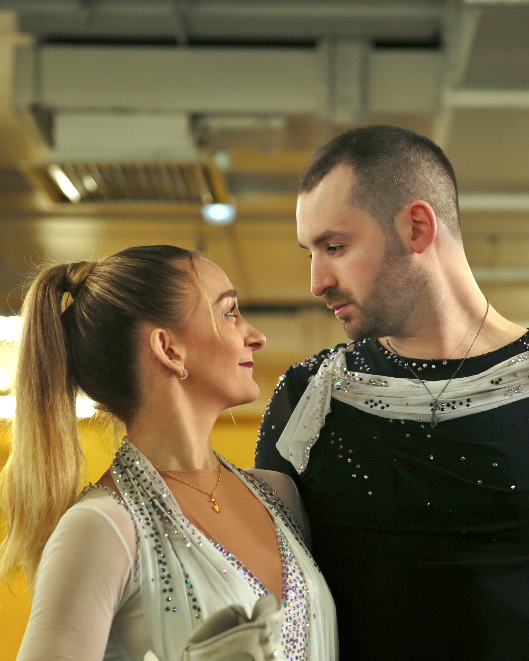 Figure skating couple in costumes looking lovingly into each other’s eyes, both wearing ice skating necklaces.