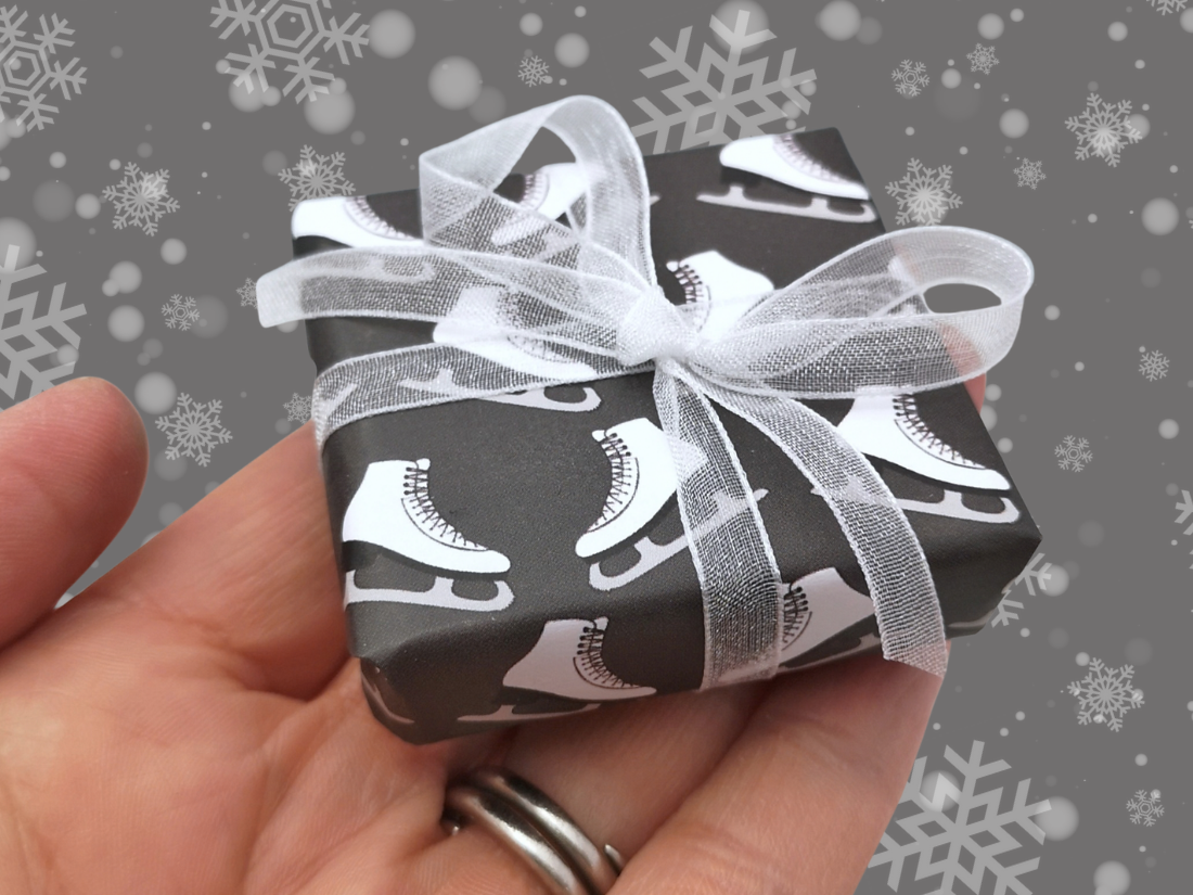 A hand holding a small gift box wrapped in black paper with a white ice skate pattern and tied with a sheer white ribbon, against a gray background with snowflake graphics.