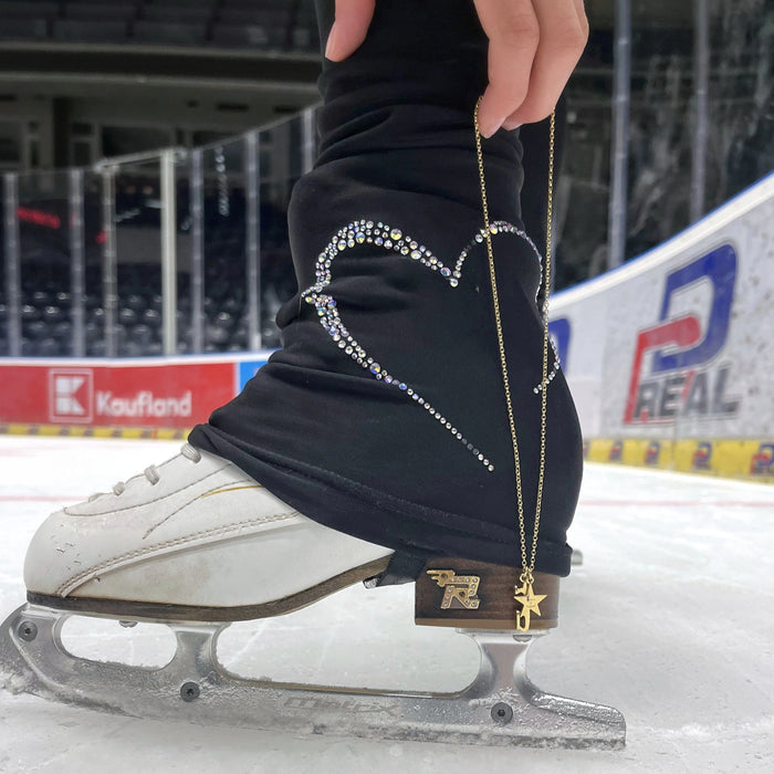 close up of an ice skating boot and blade on an ice rink and a hand holding a gold necklace with a star charm and an ice skate blade charm, near the back of the boot