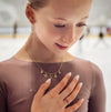 Woman wearing a gold necklace with tiny charms  and a ring, with a blurred indoor ice rink background.