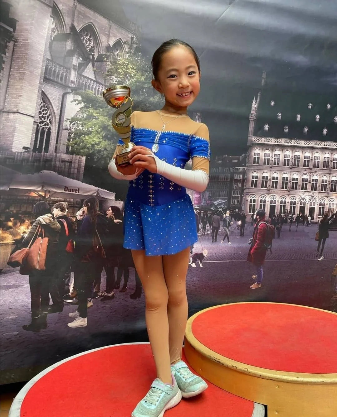 young figure skater in a blue dress on a podium holding a trophy wearing a silver ice skating necklace