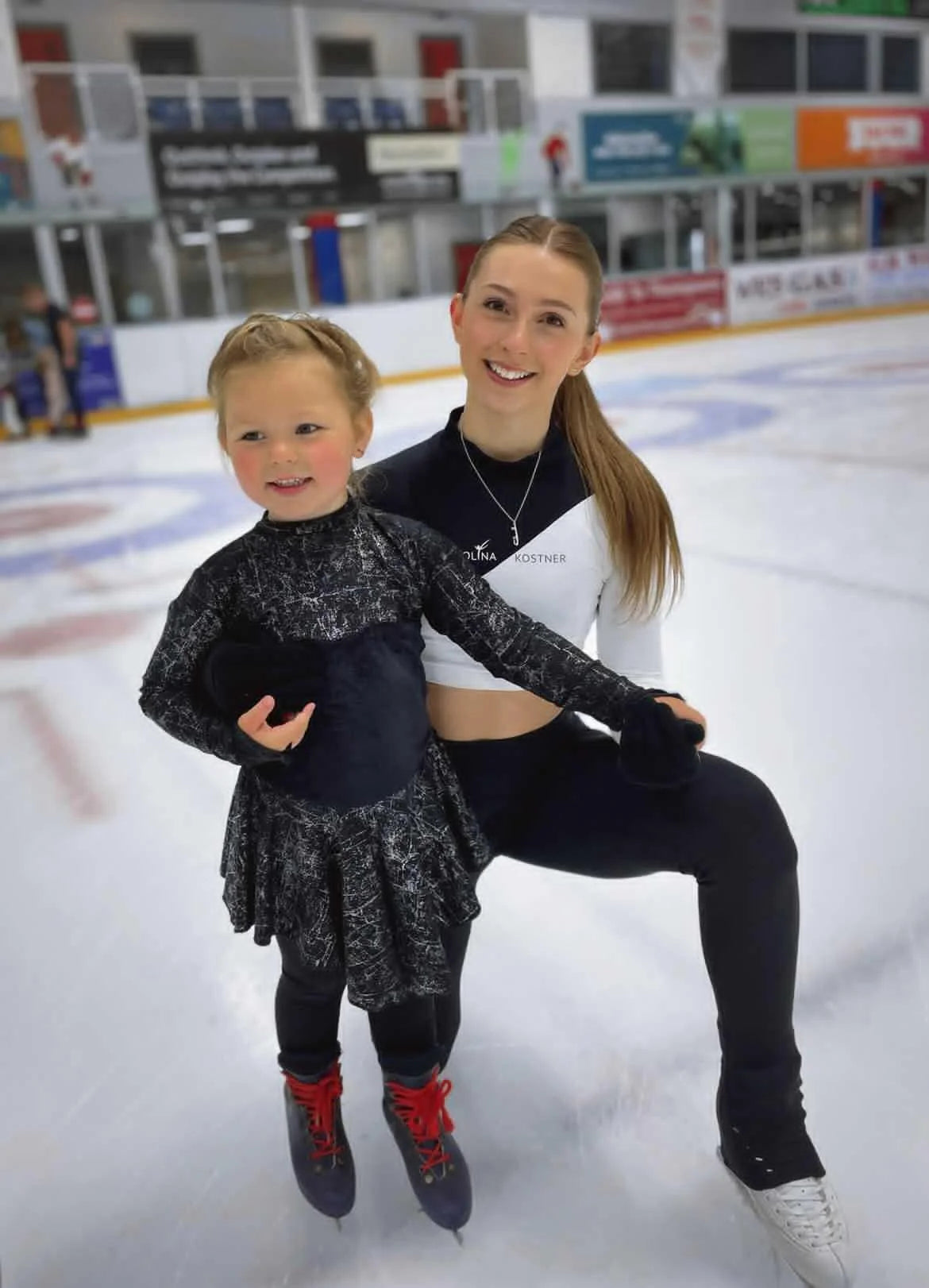 a figure skater on ice wearing a silver ice skate blade necklace holding onto a very young first-time skater in a black dress