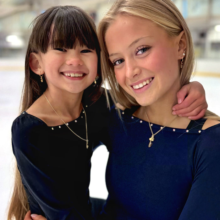 Close up of two figure skaters in dark blue and black tops on an ice rink wearing gold ice skate blade necklaces