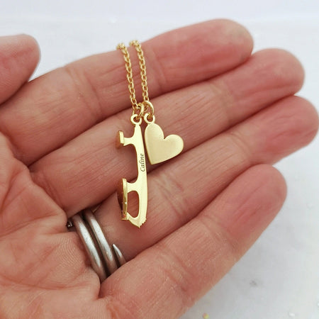 Gold necklace with a heart and an ice skate blade charm held in a hand on a white background. the blade charm is engraved with a name