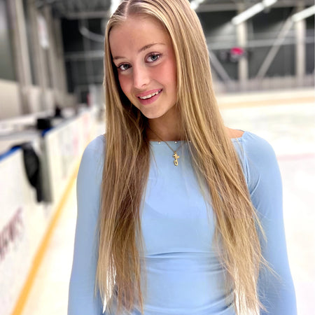 Figure skater standing on an ice rink with long blonde hair wearing a blue top and a gold ice skating necklace with a skate blade and a heart charm