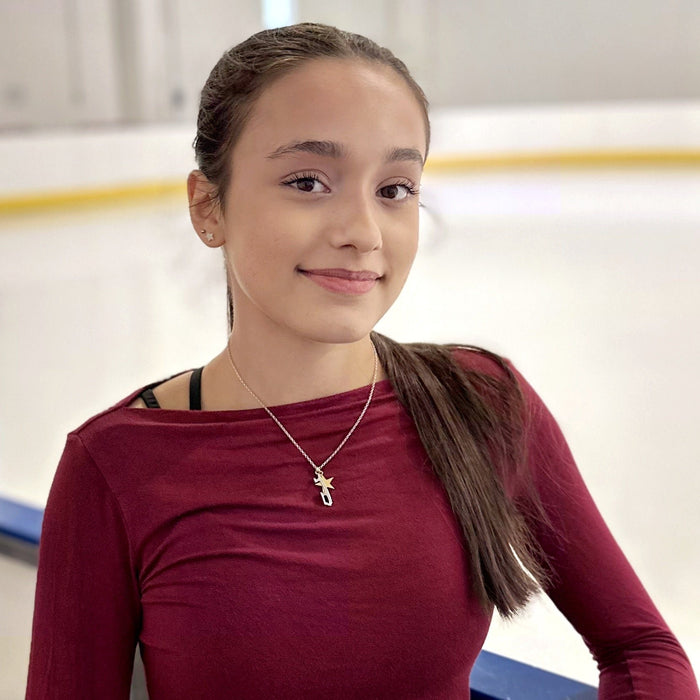 ice skater in a dark red top next to an ice rink wearing and ice skating necklace featuring a silver skate blade and a gold star