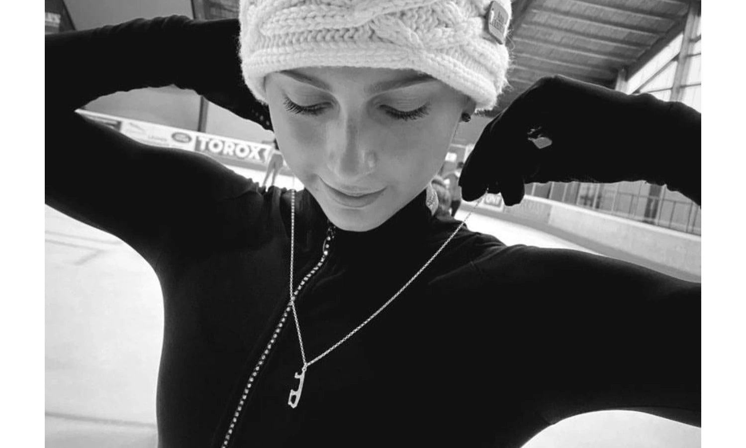 A black and white lifestyle shot of a female figure skater on ice wearing a white knit hat, black gloves, and a silver ice skating blade necklace.
