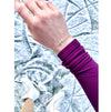 A close-up of a person's wrist wearing a delicate silver chain bracelet with a tiny ice skate blade, paired with a purple long-sleeve top and white ice skating boots, she is standing on ice.