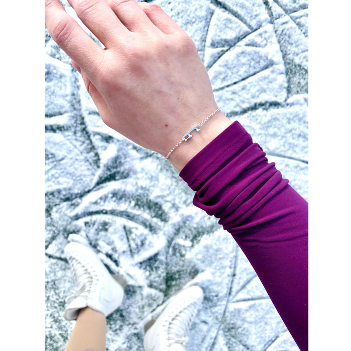 A close-up of a person's wrist wearing a delicate silver chain bracelet with a tiny ice skate blade, paired with a purple long-sleeve top and white ice skating boots, she is standing on ice.