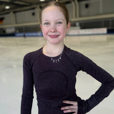 figure skater standing on ice in a dark top wearing a silver charm necklace with a star, heart, skate blade and an H