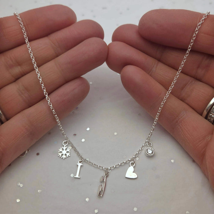 Silver charm necklace with charms held between two hands on a white background, the charms are a snowflake, letter I, skate blade, heart and a sparkly stone