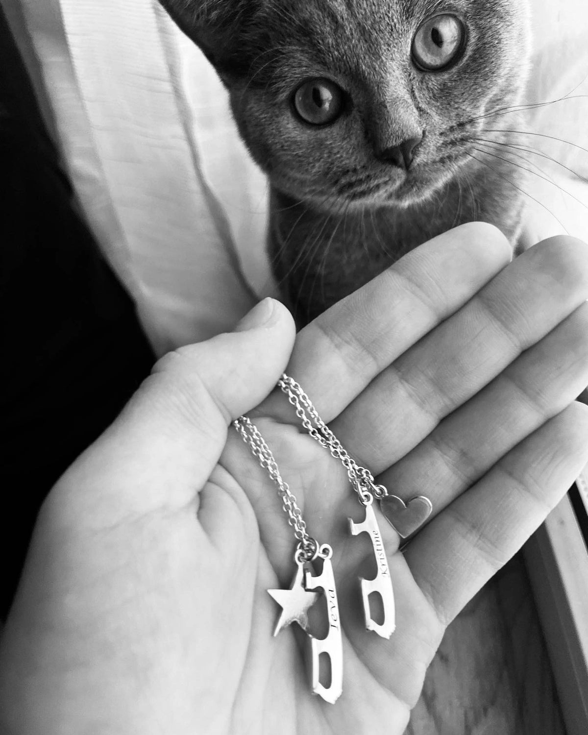 close up image of a hand holding two personalised ice skating necklaces with a heart and a star being watched by a cute cat